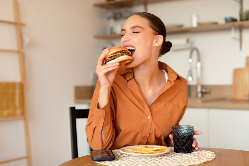 Junk food. Lady eating tasty burger, holding and biting delicious unhealthy fastfood, sitting at table in kitchen interior, free space, banner