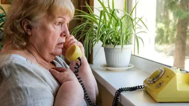 Photo of Old woman receives phone call from a scammer. Frightened grandmother talking