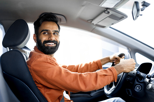 New Automobile. Cheerful Indian Car Owner Guy Sitting In Driver's Seat In Vehicle, Smiling At Camera. Man Holding Hands On Steering Wheel Posing In Auto. Transportation Offer