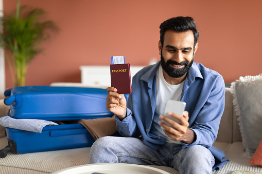 Travel Application. Happy young indian man using smartphone to book vacation online, holding international passport and plane tickets, sitting on couch near suitcase at home