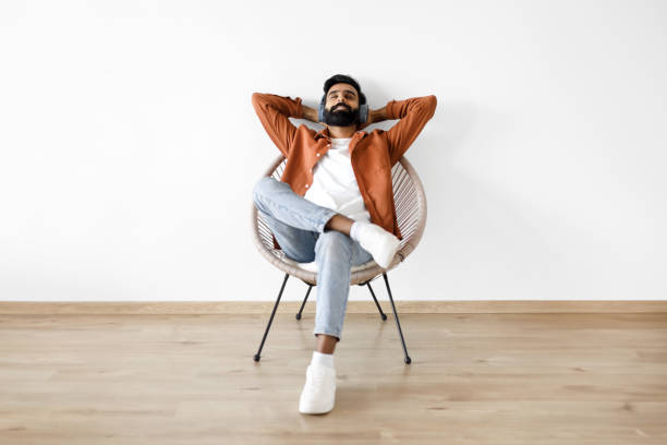 jeune homme indien détendu avec des écouteurs au repos en écoutant de la musique à l’intérieur - armchair comfortable relaxation headphones photos et images de collection