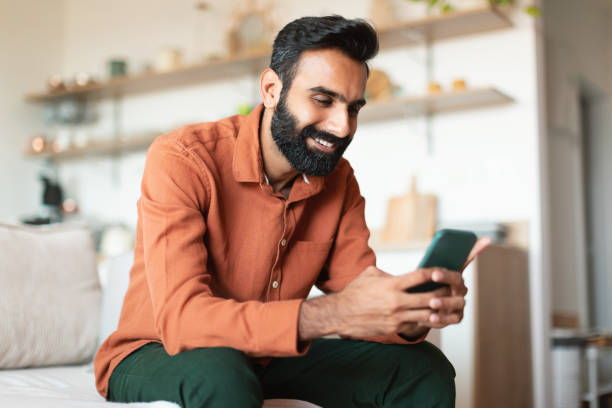 istock Cheerful Indian bearded man messaging on cell phone sitting indoor 1777780275