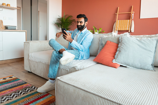 Smiling Indian guy enjoying online leisure using mobile phone, texting and communicating by cellphone at home, sitting on sofa and relaxing on weekend. Empty space for text. Gadgets and internet