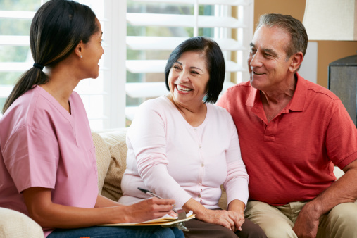 Nurse Making Notes During Home Visit With Senior Couple Talking To Each Other Smiling