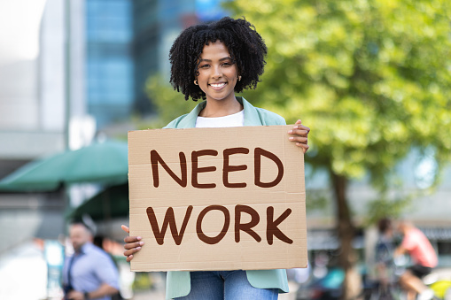 Unemployment concept. Well-motivated smiling pretty curly young black woman in smart casual outfit standing on the street, jobless african american lady showing need work placard, copy space