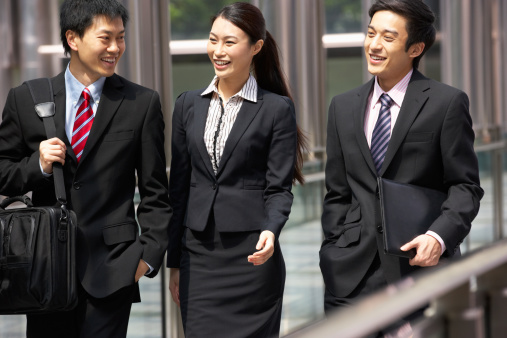 Three Business Colleagues Having Discussion Whilst Walking Outside Office Holding Briefcase And Documents Smiling