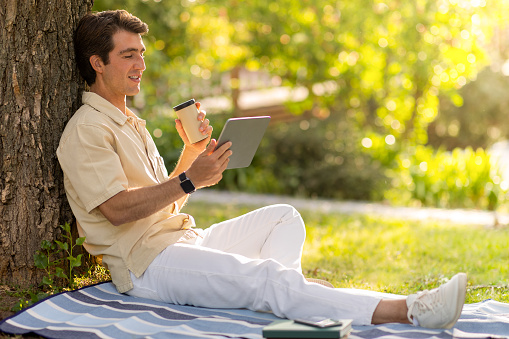 Happy handsome guy wearing casual outfit sitting under tree at public park, using digital tablet, copy space. Millennial man resting outdoors, have video call with family or lover. Telecommunication