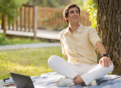 Positive attractive caucasian young man wearing casual outfit sitting on plain blanket under tree at park, have picnic, working online, using laptop, enjoying sunny summer day, looking at copy space