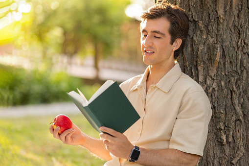 Happy handsome relaxed millennial man in beige shirt sitting under tree at public park or garden, reading book, eating apple, copy space. Relaxation, stress relief, weekend, rest concept