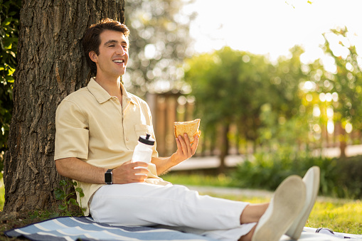 Thrilled handsome young man wearing comfy casual outfit sitting on plain blanket under tree, drinking fresh water and eating sandwich, guy have picninc at public park on sunny day, copy space