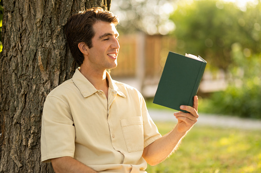 Closeup of handsome relaxed young man in beige shirt sitting under tree at public park or garden, reading book, enjoying nice story. Relaxation, stress relief, weekend, rest, summer leisure concept