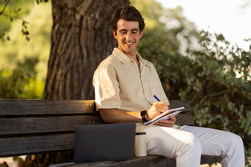 Cheerful young man attending webinar online course training while resting at park, guy sitting on bench at garden, looking at computer laptop screen, taking notes in notepad, learning new language