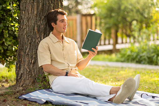 Carefree handsome relaxed young guy in casual sitting alone on blanket under tree at public park or garden, reading book. Relaxation, stress relief, weekend, rest concept, copy space