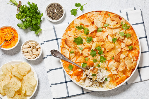 creamy cheesy chicken almond casserole topped with potato chips in baking dish with spoon on concrete table with ingredients, american recipe, flat lay