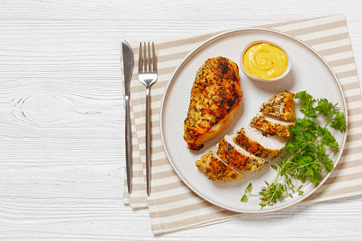 Baked Split Chicken Breasts with mustard, parsley and thyme on plate on white wooden table with cutlery, horizontal view from above, flat lay, free space