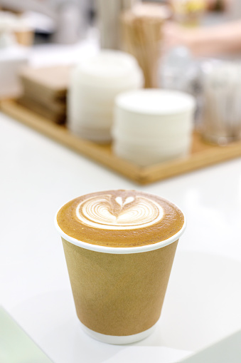 A paper cup of coffee latte placed on a barista's table, likely prepared for a customer to take away. The rich and creamy coffee is ready to be enjoyed on the go.