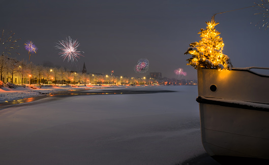 Christmas tree stands at the edge of the bow of a boat and firewors in the background. Silvester time on the Schlei. Fireworks on New Year. Promenade covered with snow in winter in Schleswig.