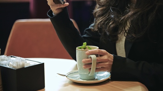 Close-up, slow motion. Beautiful Caucasian woman sitting inside of modern cafe with a lot of green plants, drinking hot tea with mint with pleasure, smiling and scrolling her smartphone.