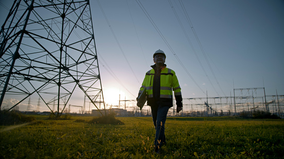 In the ambitious glow of sunset,an electrical engineer walks with determination,holding a laptop at the power station. A silhouette against the fading light,symbolizing the relentless pursuit of excellence in the electrical field