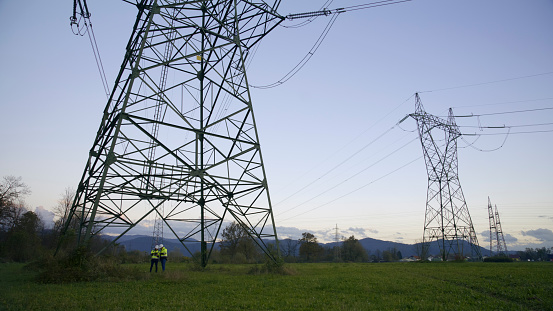 Electrical engineers stand beneath an electricity pylon on a grassy field at the power station. A representation of teamwork and expertise in the ever-evolving field of electrical engineering