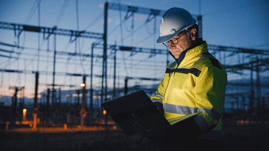 In the twilight's dim glow,a dedicated mature male electrical engineer works diligently on his laptop at a dark power plant,embodying experience and commitment in the industrial twilight