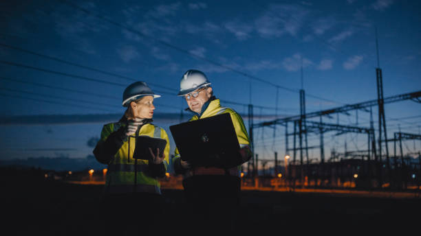 Electrical Engineers Discussing Over Laptop And Digital Tablet While Standing At Dark Power Station During Night At a dark power station in the still of night,electrical engineers engage in discussions over a laptop and digital tablet,blending technology seamlessly in their collaborative work authority stock pictures, royalty-free photos & images