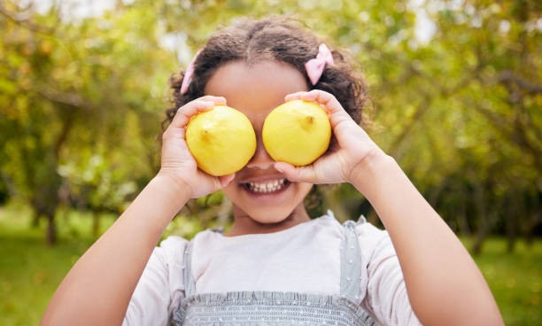 limão nos olhos, sorriso e menina na fazenda para a colheita, agricultura sustentável e crescimento na natureza. agricultura, infância e criança feliz com frutas para nutrição, alimentação saudável e alimentos orgânicos na horta - arms raised people orange blue - fotografias e filmes do acervo