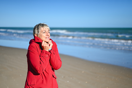 front view portrait of a mature woman with blond hair in red raincoat enjoying the sun on the beach in autumn.