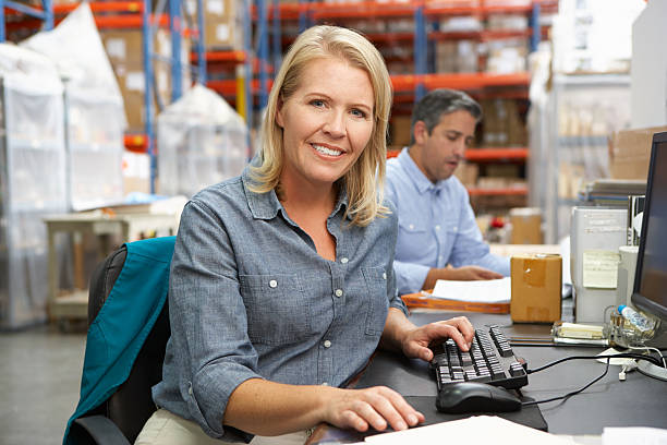 Businesswoman Working At Desk In Warehouse Businesswoman Working At Desk In Warehouse Smiling To Camera warehouse office stock pictures, royalty-free photos & images