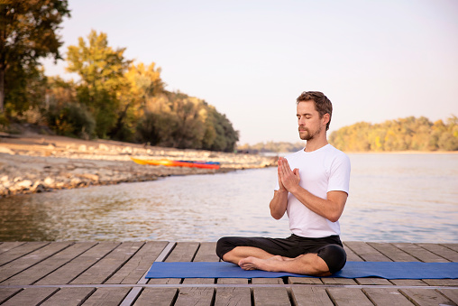 Full length of caucasian man practicing yoga on pier by lake. Mature man doing lotus position and holding hand on prayer position.