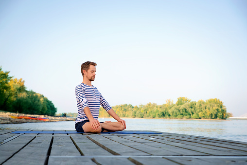 Full length of caucasian man doing yoga on pier by lake. Mature man practicing lotus position and relaxing.