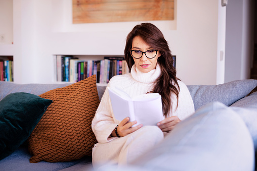 Shot of smiling woman wearing casual clothes while relaxing on the sofa at home and reading a book.