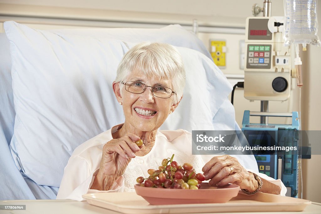 Senior Female Patient Eating Grapes In Hospital Bed Senior Female Patient Eating Grapes In Hospital Bed Smiling At Camera 80-89 Years Stock Photo