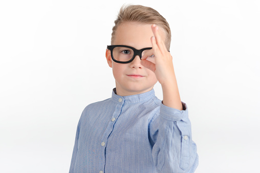 Cute caucasian boy with eyeglasses in blue shirt shows gesture ok or zero.Photo studio white bright wall.