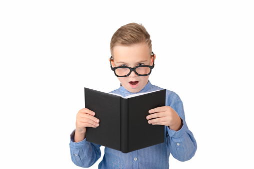 Opened mouth schoolboy reads book and wearing glasses while standing in the studio, isolated over white