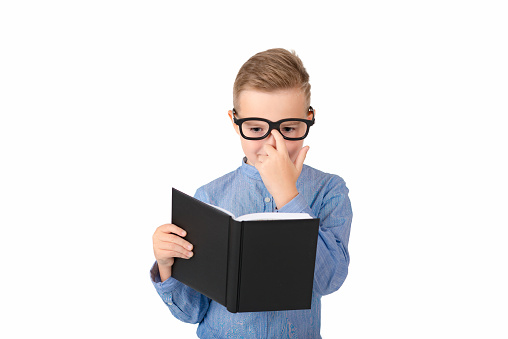 Cute dreamy schoolboy reads book and wearing glasses while standing in the studio, isolated over white