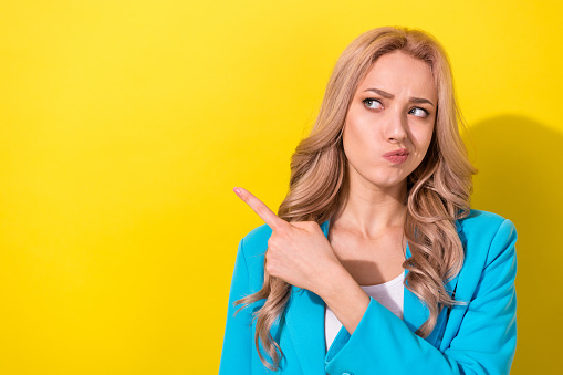 Portrait of pretty minded girl look direct finger empty space hesitate isolated on yellow color background.