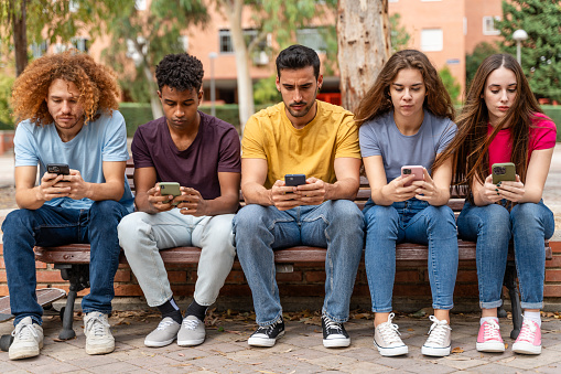 Group of young multiethnic students sitting on bench and using smartphones