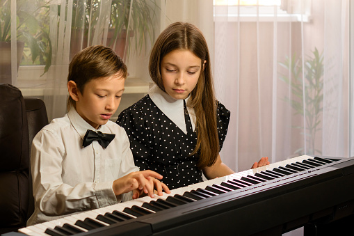 Dressed in formal attire, a young brother and sister concentrate on playing a piano duet in their living room
