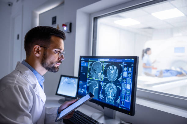Doctor examining X-ray images in MRI control room. Doctor examining X-ray images on display in control room while in background nurse preparing the patient for new MRI scanning test. doctor head stock pictures, royalty-free photos & images