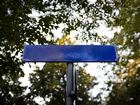 Empty blue road name sign template. Metal plate from a low angle view. The copy space can be used as a mockup for street sign in Germany. Green leaves of big trees are in the background.