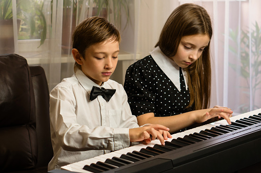 A boy and girl, dressed smartly, share a moment of music practice on a home piano.