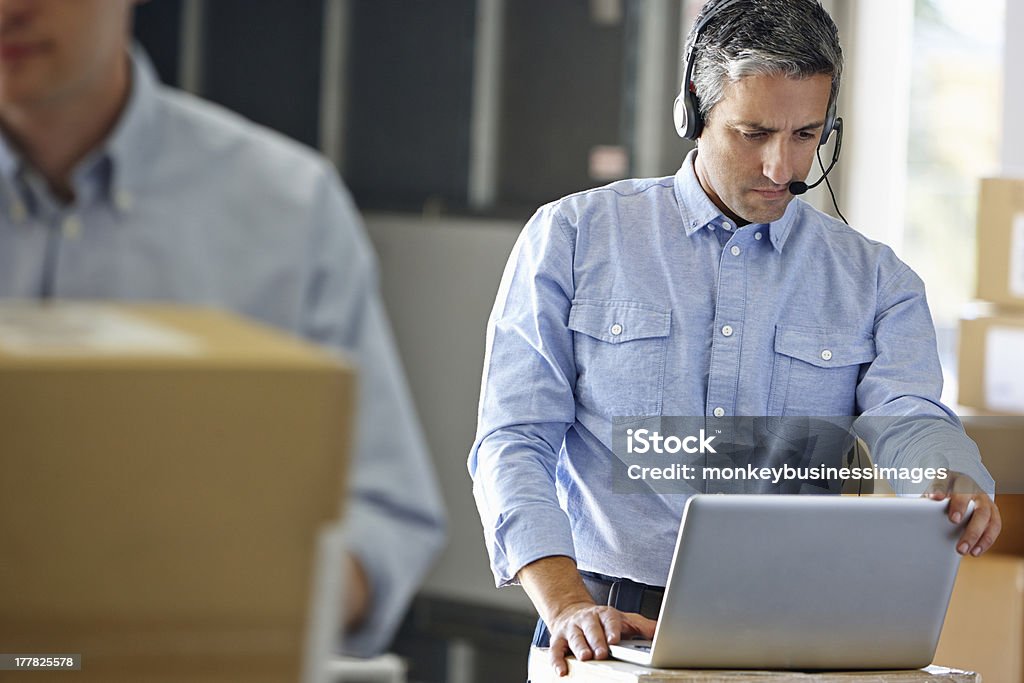 A manager using a headset and laptop Manager Using Headset In Distribution Warehouse Working On Laptop Dispatcher Stock Photo