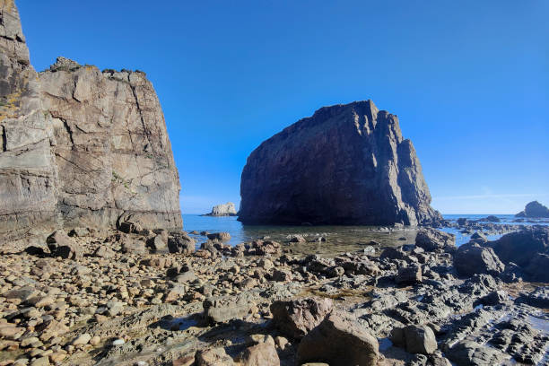 una spiaggia rocciosa con una grande formazione rocciosa frastagliata sotto un cielo azzurro limpido, con piccole rocce e massi sparsi intorno - beauty in nature cloud rocky coastline rock foto e immagini stock