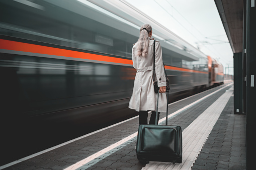 Dynamic shot of one mature adult woman with long hair, suitcase, dressed casual business walking along railway platform, rear view, train in motion blure