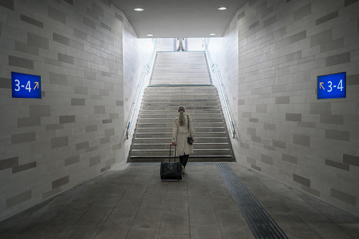 rear view of one business woman with long hair, trench coat and suitcase trolley walking to staircase leading to platform in railroad station