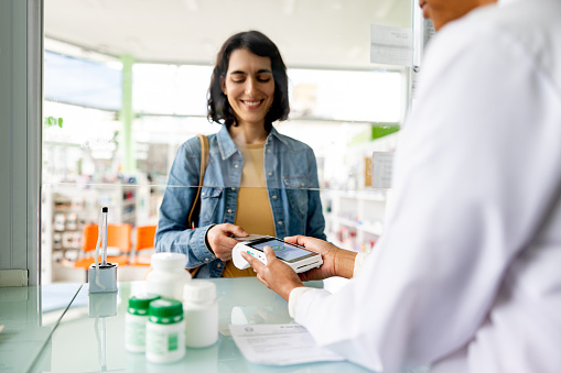Happy Latin American woman paying by card while buying medicines at the pharmacy - tap to pay concepts