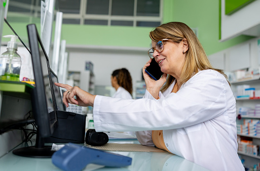 Latin American pharmacist taking a delivery order on the phone at the drugstore while checking availability on the computer - healthcare business concepts