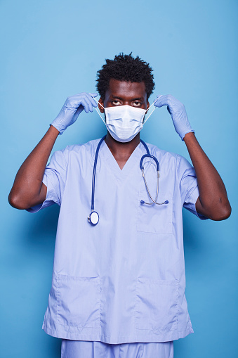 Portrait of a medical specialist putting on face mask to defend against the covid 19 epidemic. Man with blue scrubs, stethoscope, and gloves staring at camera, working as a nurse.