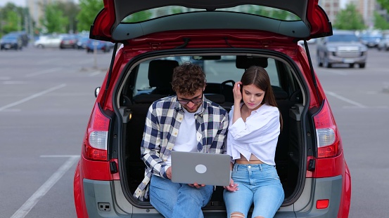 Happy young people sitting on the trunk car outdoors while using laptop, looking on screen. Transportation, technology, trip, travel and lifestyle concept. Slow motion